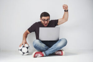 Homem com notebook comemorando vitória em 1xBet com uma bola de futebol.